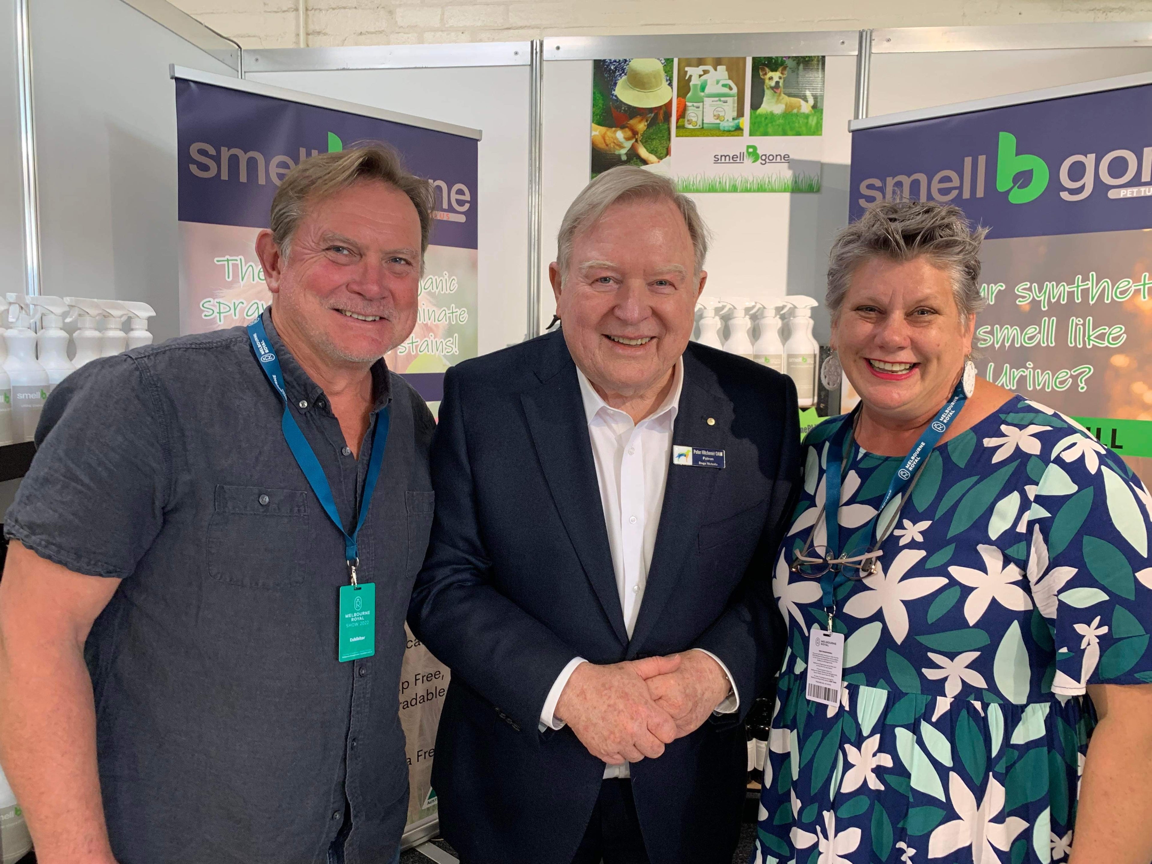 Three smiling adults stand together at a booth with “smell b gone” banners and products displayed in the background. Two are casually dressed, and the man in the center wears a dark suit.